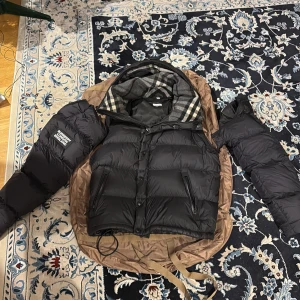 Svart pufferjacka från Burberry M - Säljer en svart pufferjacka från Burberry i storlek M. Jackan har avtagbara ärmar så den kan användas som väst, huva och klassiskt rutigt foder. Stora fickor med dragkedja och Burberry-patch på ena ärmen. Perfekt för kalla dagar och riktigt snygg streetstil.