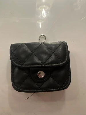 Svart ”miniväska”  - Liten svart plånbok med quiltad design och silvrig knapp framtill. Har en metallögla upptill för att fästa i väska eller nyckelring. Perfekt för kort eller småsaker när du vill resa lätt. 