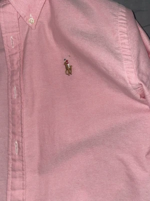 Rosa skjorta från Ralph Lauren - Säljer en rosa skjorta från Ralph Lauren i slim fit med klassisk button-down krage och broderad logga på bröstet. Skjortan har långa ärmar och vita knappar framtill. Perfekt för dig som vill ha en stilren och tidlös look.