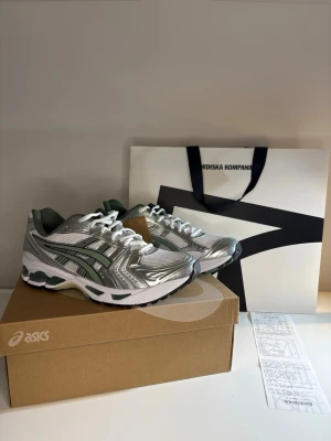 Asics sneakers i silver och vitt - Säljer ett par riktigt snygga Asics sneakers i silver, vitt och grönt. Skorna har meshpaneler för bra andningsförmåga, detaljer i syntet och klassisk snörning. Sulan är vit med svarta och gröna inslag och ger grym komfort till vardags eller träning.