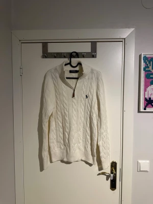 Vit kabelstickad polotröja Polo Ralph Lauren - Kabelstickad vit polotröja från Polo Ralph Lauren med half zip och ribbad krage. Klassisk logga broderad på bröstet. Tröjan är långärmad och har en chunky, mysig känsla som passar perfekt till kyligare dagar.