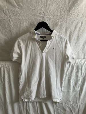 Vit pikétröja från Tommy Hilfiger - Klassisk vit pikétröja från Tommy Hilfiger med liten broderad logga på bröstet. Tröjan har korta ärmar, krage och knappar framtill. Tillverkad i mjuk bomull som känns skön mot huden. Perfekt till jeans eller shorts för en clean look.