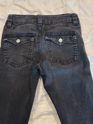 Bootcut svarta jeans med ficklock - Säljer ett par svarta bootcut jeans med snygg tvätt och coola ficklock bak med silverknappar. Jeansen har klassisk femficksmodell, bälteshällor och är i stretchigt denim. Perfekta för dig som gillar en retrovibe och vill ha ett par jeans som sticker ut lite extra.