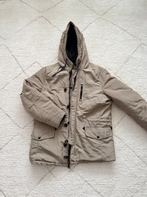 Beige parkajacka från Mango Man XL - Säljer en beige parkajacka från Mango Man i storlek XL. Jackan har huva med snörning, flera fickor med knappar, dragkedja och knappar framtill samt quiltat svart innerfoder. Perfekt för kalla dagar och riktigt snygg till streetwear.