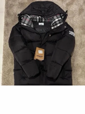 Svart pufferjacka från Burberry - Säljer en svart pufferjacka från Burberry med stor huva och klassiskt rutigt foder i svart, vitt och rött. Jackan har en tydlig Burberry London England-patch på ärmen och dragkedja framtill. Perfekt för kalla dagar och riktigt snygg streetstil.