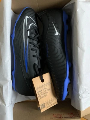 Nike Phantom svart/blå fotbollsskor - Helt nya och oanvända Nike Phantom fotbollsskor i svart med blå sula och silverfärgade detaljer. Snyggt mönster på ovansidan och klassisk snörning. Skorna har rund tå och platt sula med dobbar för gräsplan. Tillverkade i syntetmaterial och designade för att ge bra grepp och kontroll på planen. Nypris: 757 mitt pris: 350. Priset kan diskuteras vid snabb affär 
