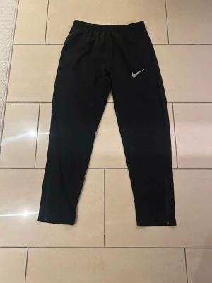 Svarta träningsbyxor från Nike - Svarta träningsbyxor från Nike med smal passform och elastisk midja. Byxorna har en diskret Nike-logga på ena benet och dragkedjor vid bensluten för extra funktionalitet. Perfekta för sport eller chill. 