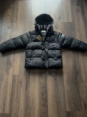 Svart pufferjacka från Burberry - Säljer en stilren svart pufferjacka från Burberry med avtagbara ärmar, vilket gör att den även kan bäras som väst. Jackan har en avtagbar huva fodrad med Burberrys klassiska rutmönster, dragkedja framtill samt flera praktiska fickor.  Ett mycket varmt och bekvämt plagg – perfekt för kalla dagar och flexibel nog att användas i olika väderförhållanden. Sickar samma dag som du köper!