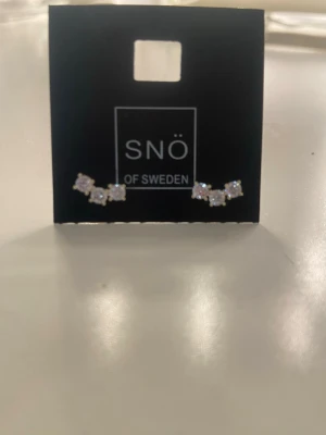 Örhängen med stenar från SNÖ of Sweden - Säljer ett par små örhängen från SNÖ of Sweden med tre gnistrande klara stenar i rad. Örhängena har en böjd form som följer örsnibben och är i silverfärg. Perfekta för att ge lite extra bling till din look.