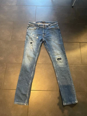 Blå slitna jeans från Jack & Jones - Säljer ett par blå jeans från Jack & Jones med flera slitningar och hål på benen för en riktigt edgy look. Klassisk femficksmodell med normal passform och raka ben. Perfekta för dig som gillar en avslappnad och trendig stil.