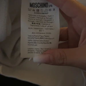 Vit hoodie från Moschino Couture - Snygg vit hoodie från Moschino Couture med svart logga och tryck på bröstet. Tröjan har huva med snörning och är tillverkad i 100% bomull för en skön och mjuk känsla. Perfekt för dig som gillar stilrena och exklusiva plagg.
