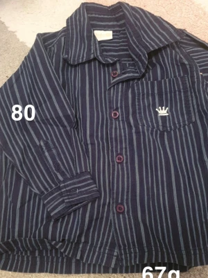 Blå randig skjorta med ficka - Mörkblå skjorta från Lindex med tunna ljusblå ränder och en bröstficka med broderad krona. Skjortan har långa ärmar, klassisk krage och knappar framtill. Perfekt för en cool och avslappnad stil.🌸 Fint använt skick. Storlek 80