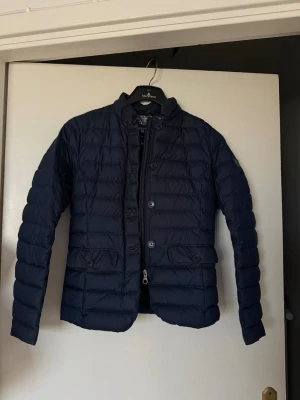 Mörkblå quiltad pufferjacka från Gant - Säljer en mörkblå quiltad pufferjacka från Gant i storlek XS. Jackan har snygga knappar och dragkedja framtill samt två fickor med lock. Insidan har ett unikt mönster i blått, vitt och guld. Perfekt för dig som vill ha en lätt och stilren jacka med coola detaljer. Ny pris 2000 kr