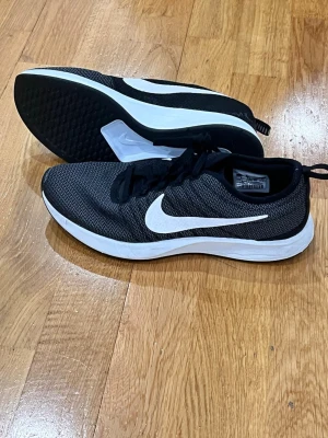 Svarta Nike sneakers med vit sula39 - Säljer ett par stilrena svarta Nike sneakers med vit sula och stor vit swoosh på sidan. Ovandelen är i mesh-liknande syntetmaterial för bra andningsförmåga och komfort. Perfekta för träning eller vardagsbruk. Klassisk rund tå och snörning.