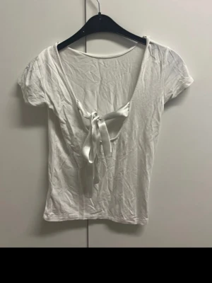 Vit topp med knyt bak - Vit topp med korta ärmar och djup v-ringning framtill som knyts med ett brett band. Tunt och mjukt material som känns skönt mot huden. Enkel och stilren design som passar till många olika outfits. Knappt använd felfri!