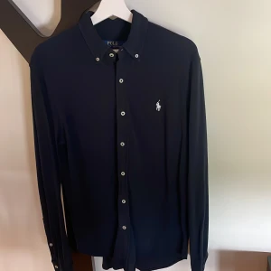 Mörkblå skjorta från Polo Ralph Lauren - Snygg mörkblå skjorta från Polo Ralph Lauren i featherweight mesh. Klassisk design med vit broderad logga på bröstet, knappar framtill och långärmad modell. Perfekt för dig som gillar stilrena och tidlösa plagg med premiumkänsla. Passar dig som är omkring 185cm lång.