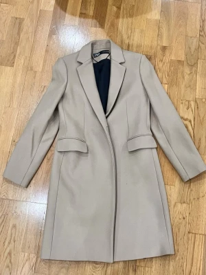 Beige och svart kappa från Zara - Två stilrena kappor från Zara i klassisk modell. Ena är beige och den andra svart, båda med långa ärmar, krage och fickor framtill. Perfekta för höst och vinter, tillverkade i ullblandning för extra värme. Snygg passform och tidlös design. 500kr styck 800 för båda