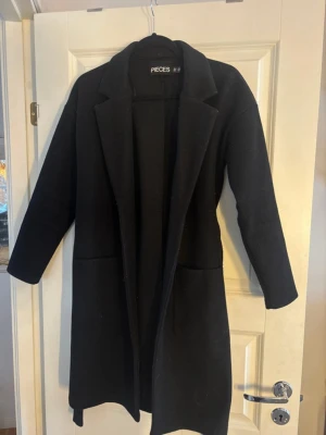 Svart kappa från Pieces med bälte XS - Svart lång kappa från Pieces i storlek XS. Klassisk design med krage, två stora fickor framtill och bälte i midjan. Jackan är i ett mjukt material och har lång ärm. Perfekt för höst och vinter när du vill ha en stilren look.