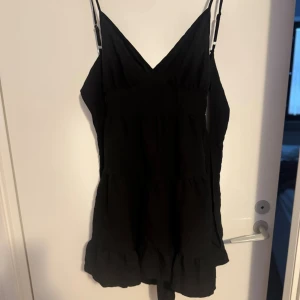 Svart kort klänning med volang - Säljer en svart kort klänning med smala axelband och v-ringning. Klänningen har volangdetaljer nedtill och en öppen rygg som ger en snygg touch. Man knyter även klänningen bak i ryggen. Bara använd 1 gång så i väldigt bra skick!