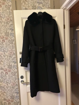 Svart kappa från Meotine med bälte - Lång svart kappa från Meotine med bred fuskpäls-krage (som är avtagbar) och bälte i midjan. Ärmarna har snygga spännen och kappan har en klassisk, rak passform. Lite noppig längs med på ena sidan (se bild).