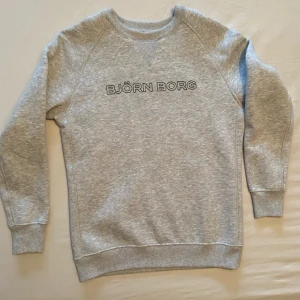 Grå sweatshirt från Björn Borg - Klassisk grå sweatshirt från Björn Borg med rund hals och svart broderad logga på bröstet. Tröjan har långa ärmar, ribbade muddar och är tillverkad i mjuk bomullsblandning. Perfekt för en avslappnad och sportig stil.