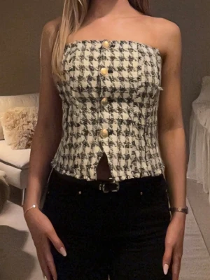 Rutig axelbandslös tubtopp med knappar - Säljer en snygg axelbandslös tubtopp i svartvitt rutigt tyg. Toppen har dekorativa guldfärgade knappar framtill och en liten slits nedtill.