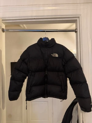 Svart dunjacka från The North Face - Snygg svart dunjacka från The North Face med hög krage och dragkedja framtill. Jackan har en puffig design, logga på bröstet och ryggen samt justerbar dragsko nedtill. 