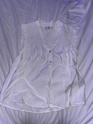 Vit ärmlös blus med smockdetaljer - Söt vit blus med smock vid axlar och rygg, v-ringning och tre knappar framtill i storlek small. Blusen är ärmlös och har en lätt, luftig känsla med volangkant vid halsen. Perfekt för en fräsch och stilren look.