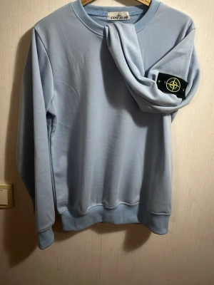 Ljusblå sweatshirt från Stone Island - Säljer en ljusblå sweatshirt från Stone Island med den klassiska logotypen på vänster ärm. Tröjan har rund halsringning, ribbade muddar och är tillverkad i mjukt bomullsmaterial. Perfekt för en clean och stilren look.