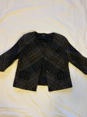 Rutig kort kappa från Lindex - Snygg kort kappa i brun rutigt. Modellen har trekvartsärmar, rund halsringning och tre stora knappar framtill. Två fickor framtill och helfodrad insida. Perfekt för en trendig och stilren look.