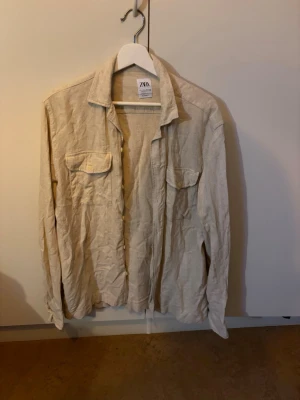 Beige skjorta från Zara - Luftig och avslappnad beige skjorta från Zara i linne. Skjortan har två bröstfickor med lock, klassisk krage och långa ärmar. Perfekt för en chill och stilren look.