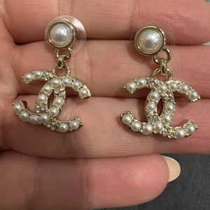 Chanel örhängen med pärlor och strass - Supersnygga Chanel örhängen i guld med ikoniska CC-loggan, dekorerade med vita pärlor och glittrande strass. Örhängena har en rund pärla upptill och hängande logga, perfekt för att lyfta din look med lyxig vibe.