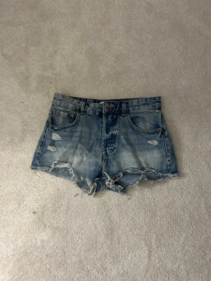 Blå denimshorts med slitningar - Säljer ett par blå denimshorts med råa kanter och slitna detaljer framtill. Shortsen har klassisk femficksdesign, bälteshällor och knappgylf. Perfekta för varma dagar och en avslappnad look.