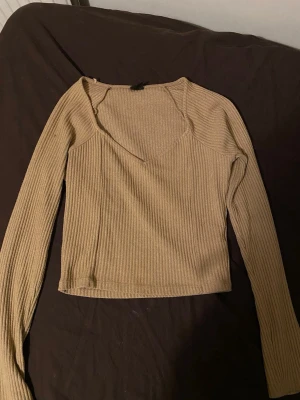 Beige ribbad långärmad topp - Trendig beige långärmad topp med ribbad struktur och djup rundad halsringning. Croppad modell som passar perfekt till höga jeans eller kjol. Tillverkad i mjukt stretchigt material för en bekväm passform.