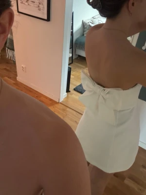 Vit axelbandslös kort klänning - Säljer en vit axelbandslös kort klänning med en stor rosett i ryggen. Klänningen har en enkel, elegant design och är perfekt. Materialet känns slätt och stadigt, och modellen är figurnära upptill med lätt utsvängd kjol. perfekt för studenten och andra högtider. använt få antal gånger! 