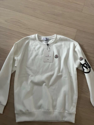 Vit sweatshirt från Moncler - Snygg vit sweatshirt från Moncler med rund halsringning och ribbade muddar. Tröjan har Moncler-logga på bröstet och ärmen samt en stor svart grafisk detalj på ena ärmen. Perfekt för en clean och trendig look.