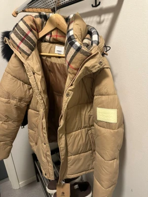 Beige pufferjacka från Burberry - Snygg beige pufferjacka från Burberry med klassiskt rutigt foder i huvan och stor patch med Burberry London England på ärmen. Jackan har dragkedja, fickor och en rejäl huva med fluffig känsla. Perfekt för kalla vinterdagar och riktigt trendig look.