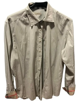 Beige skjorta från Burberry - Klassisk beige skjorta från Burberry med lång ärm och knappar framtill. Skjortan har diskret broderad logga på bröstet och ikoniskt rutigt tyg på insidan av manschetterna. Perfekt för en stilren look.