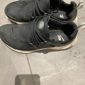 Nike Presto svarta sneakers - Svarta Nike Presto sneakers med mesh-ovandel och elastiska snören. Skorna har en vit sula och diskret Nike-logga på sidan samt på plösen. Låg modell med sportig och clean design, perfekt för en streetwear-look.