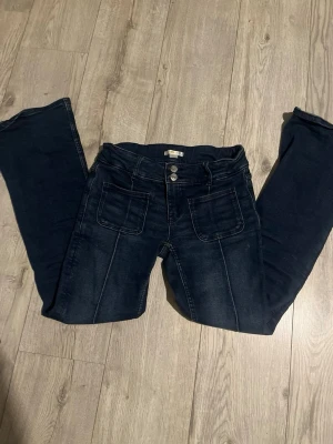 Mörkblå utsvängda jeans Gina Tricot - Säljer ett par mörkblå jeans från Gina Tricot med utsvängda ben och dubbla knappar i midjan. Jeansen har stora fickor framtill och markerade sömmar som ger en snygg retrovibe. Perfekta för dig som gillar en trendig och avslappnad stil.