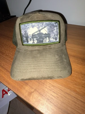 Olivgrön truckerkeps med tryck - Säljer en olivgrön truckerkeps med mesh på sidorna och ett stort tygmärke framtill med svartvitt motiv av ett piano och människor. Kepsen har böjd skärm och justerbar baksida. Perfekt för dig som gillar unik streetwear och coola detaljer.