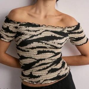 Zebramönstrad smockad topp - Trendig zebramönstrad topp med smockad struktur och korta ärmar. Toppen har en off shoulder-modell och vågig kant nertill och vid ärmslut. Färgerna är svart och beige, vilket ger en cool kontrast. Perfekt för dig som gillar djurmönster och vill sticka ut. Köpt från JDY / Nelly, aldrig använt lappen sitter kvar 🦓🤍