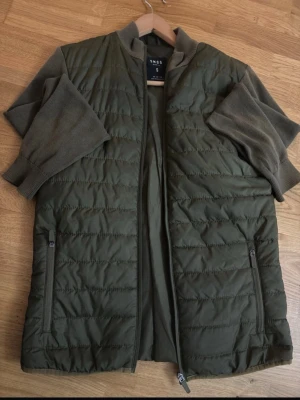 Olivgrön dunväst från Smog - Snygg olivgrön dunväst från Smog med dragkedja framtill och två praktiska fickor med dragkedja. Västen har en quiltad design och står ut med sin stilrena look. Perfekt för lager på lager och enkel att matcha med olika outfits.