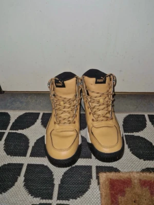 Beige kängor från Puma - Snygga beige kängor från Puma med högt skaft och svarta detaljer. Skorna har kraftig sula, snörning framtill och vadderad krage för extra komfort. Tillverkade i slitstarkt material som passar perfekt till streetwear och vardagsstil.