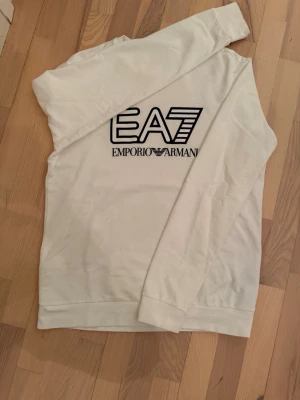 Vit EA7 Emporio Armani sweatshirt/tröja - Vit sweatshirt från EA7 Emporio Armani med stor svart logga framtill. Tröjan har rund halsringning och långa ärmar. Perfekt för en clean och stilren look. Materialet känns mjukt och bekvämt mot huden. Den är i storlek XL men passar mig som har L. Har även en jätte liten fläck vid armen men inget som syns mycket annars i bra skick.