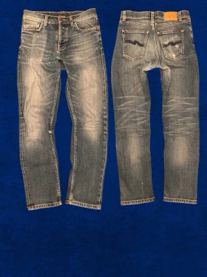 Blå slitna jeans från Nudie Jeans - Säljer ett par blå jeans från Nudie Jeans med snyggt slitna detaljer och klassisk femficksmodell. Jeansen har raka ben, kontrastsömmar och den ikoniska vågstickningen på bakfickorna. Tillverkade i denim med en cool tvättad look.