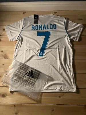 Real Madrid Ronaldo #7 fotbolls tröja - Vit Real Madrid matchtröja från Adidas med blå detaljer, Ronaldo och nummer 7 på ryggen. Framsidan har klubbmärke, Adidas-logga och Fly Emirates-tryck. Korta ärmar och klassisk V-ringning. Perfekt för fotbollsfans! Lite mindre än vanliga size M tröjor