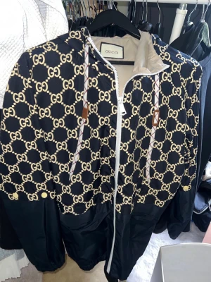Gucci vindjacka med GG-mönster - Svart vindjacka från Gucci med beige GG-monogram över hela ovandelen. Jackan har huva med dragsko, vita detaljer och dragkedja framtill. Materialet är lätt och glansigt, perfekt för säsongens skiftande väder.