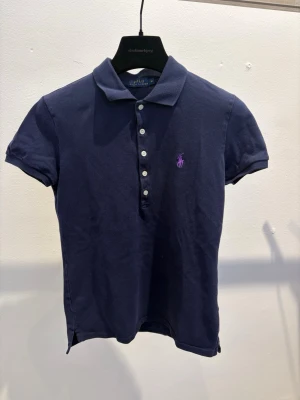 Marinblå pikétopp från Ralph Lauren - Klassisk marinblå pikétopp från Ralph Lauren med korta ärmar och krage. Toppens knäppning framtill har vita knappar och den ikoniska loggan i lila brodyr på bröstet. Perfekt för en stilren look.