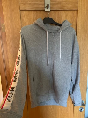 Grå hoodie från Moschino med dragkedja - Grå hoodie från Moschino med vit dragkedja och huva. Snygga vita band med Moschino-logga och röda detaljer längs ärmarna. Mjuk bomullskänsla och fickor framtill. 